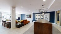 This contemporary single-floor amenity lounge is designed with a refined, modern aesthetic that balances comfort and functionality. The open-plan layout is anchored by large-format tile flooring throughout, lending the space a clean, cohesive feel. A generously sized seating area serves as the heart of the lounge, featuring a rich cognac leather sectional sofa accented with colorful throw pillows, a sleek coffee table, and a coordinating blue accent chair arranged atop a boldly patterned area rug. A wall-mounted flat-screen television is set against a white subway tile feature wall, adding a media-focused focal point to the lounge area.

Adjacent to the seating zone, a substantial dark walnut communal table with gold metal bar stools provides a dedicated space for gathering or collaborative work, illuminated overhead by striking geometric pendant light fixtures. A curated gallery wall of black-and-white framed photographs adds a local, artistic touch against a deep navy accent wall.

A wet bar area extends further into the space, while glass-panel doors connect seamlessly to a bright, adjacent room. Full-height glass doors open directly onto a generous outdoor courtyard patio, complete with concrete pavers, decorative planters, and views of the surrounding multi-story building, inviting an effortless indoor-outdoor lifestyle.