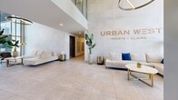 Located at 17 Place de la Triade in Pointe-Claire, QC, this building lobby offers a striking first impression through its contemporary architectural design. The entrance is defined by expansive floor-to-ceiling glass panels flanking double doors, flooding the space with abundant natural light and providing clear views to the outdoors. Soaring double-height ceilings amplify the sense of openness, while clean white walls and large-format neutral stone-effect floor tiles create a polished, cohesive aesthetic throughout.

A thoughtfully arranged lounge area greets visitors with cream-toned sofas, a round marble-topped coffee table with gold accents, and a sculptural circular pendant light overhead. Lush indoor plants add a touch of warmth and organic texture to the otherwise sleek interior. A feature wall bearing the "Urban W – Pointe-Claire" branding anchors the reception zone with a refined, upscale identity.

The hallway section connects the main lobby to the building's vertical circulation, featuring two stainless steel elevators and a wall of individual resident mailboxes. Recessed ceiling lighting guides residents along the corridor, which maintains the same large-format tile flooring for visual continuity. Overall, the lobby presents a welcoming, functional, and well-appointed space designed to reflect the quality of the building it serves.