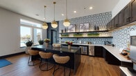 This thoughtfully designed single-floor amenity space blends contemporary style with inviting comfort across two distinct zones. The kitchen and bar area serves as a visual centerpiece, distinguished by a striking geometric-patterned tile backsplash in blue, grey, and white tones that extends across the full wall. Dark espresso cabinetry with open floating shelves, white countertops, and a large dark-topped island provide ample workspace and gathering space. Woven rattan barstools add a warm, textural contrast, while a pair of globe pendant lights in brushed brass cast a warm glow overhead. Stainless steel appliances and a dedicated coffee station complete the well-appointed kitchen. Rich wood-look flooring runs throughout, grounding the space with warmth. The adjacent lounge area offers a sophisticated retreat, anchored by a dramatic floor-to-ceiling dark paneled accent wall with a mounted flat-screen television. Plush sectional sofas in muted blues and creams are arranged around a marble-topped cocktail table, creating an intimate setting for relaxation or socializing. Large windows allow natural light to filter in, providing views to the exterior. Abstract framed artwork adorns the walls, adding a refined artistic touch throughout the space.