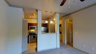 This single-floor residence offers a thoughtfully designed open-concept layout that connects the living and kitchen areas with a sense of flow and functionality. The kitchen is well-appointed with stainless steel appliances, including a gas range, microwave, and refrigerator, complemented by rich dark wood cabinetry. A raised breakfast bar with decorative pendant lighting serves as a natural dividing element between the kitchen and the adjacent living space, while white structural columns add an architectural detail that enhances the overall character of the interior. The living area features plush carpeting and is topped with a ceiling fan, contributing to a comfortable and relaxed atmosphere. Warm, ambient lighting throughout the space creates an inviting tone. An elegant arched doorway leads to an additional area, hinting at further functional spaces within the home. Built-in shelving visible through the arched passage adds a practical and decorative touch. Track lighting in the kitchen ensures the workspace is well-lit, and tile flooring provides a durable, easy-to-clean surface in the cooking area. The overall design balances practicality with tasteful architectural details, making it a welcoming and livable space.