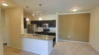 This single-floor residence offers a thoughtfully designed open-concept layout that seamlessly connects the kitchen and living areas, creating a bright and functional living environment. The kitchen is a standout feature, showcasing dark espresso cabinetry that contrasts elegantly with light granite countertops. A central island with a built-in sink and matching granite surface provides additional workspace and serves as a natural gathering point. Stainless steel appliances, including a refrigerator, over-the-range microwave, and electric range, add a sleek, modern touch. Pendant lighting hangs above the island, complemented by a track lighting fixture, ensuring the kitchen is well-lit for both cooking and entertaining. The adjacent living area transitions smoothly via neutral-toned carpeting, offering a comfortable and cohesive feel throughout. A warm, two-tone color palette featuring soft beige and olive-toned accent walls ties the space together with understated elegance. Crown molding details add a refined architectural touch. The open flow between spaces makes this residence feel welcoming and versatile, well-suited for both everyday living and hosting guests.