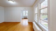 This single-floor unit offers an efficient and functional layout comprising a living room, kitchen, and bathroom. The living area is characterized by warm hardwood flooring that extends throughout the space, bright white walls, and a textured ceiling. Large multi-pane windows allow generous natural light to fill the rooms, while classic cast-iron radiators provide heating—details that speak to the building's traditional character. The open connection between the main living area and a secondary room creates a sense of continuity, with a doorway framing views through to a window-lit room beyond.

The kitchen is compact and practical, fitted with white cabinetry, a white tile backsplash, a coil-burner electric range, a refrigerator, and a stainless steel sink set into a countertop. Upper and lower cabinets provide ample storage for the space's scale.

The bathroom features a full soaking tub with a showerhead and hand-held shower attachment. The tub surround is finished in white square ceramic tile, and a stainless steel grab bar has been installed for added safety and accessibility.

Overall, the space presents a clean, no-frills aesthetic with functional finishes. The consistent hardwood flooring, natural light, and classic radiator heating lend the unit a timeless, utilitarian charm well-suited to practical everyday living.