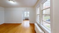 This single-floor unit offers an efficient and functional layout comprising a living room, kitchen, and bathroom. The living area is characterized by warm hardwood flooring that extends throughout the space, bright white walls, and a textured ceiling. Large multi-pane windows allow generous natural light to fill the rooms, while classic cast-iron radiators provide heating—details that speak to the building's traditional character. The open connection between the main living area and a secondary room creates a sense of continuity, with a doorway framing views through to a window-lit room beyond.

The kitchen is compact and practical, fitted with white cabinetry, a white tile backsplash, a coil-burner electric range, a refrigerator, and a stainless steel sink set into a countertop. Upper and lower cabinets provide ample storage for the space's scale.

The bathroom features a full soaking tub with a showerhead and hand-held shower attachment. The tub surround is finished in white square ceramic tile, and a stainless steel grab bar has been installed for added safety and accessibility.

Overall, the space presents a clean, no-frills aesthetic with functional finishes. The consistent hardwood flooring, natural light, and classic radiator heating lend the unit a timeless, utilitarian charm well-suited to practical everyday living.