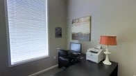 This single-floor business center is a focused, functional workspace designed to support a variety of professional needs. The room features neutral gray walls that create a calm, distraction-free environment, complemented by dark-toned flooring that adds a touch of sophistication. A large window fitted with horizontal blinds allows natural light to fill the space while offering flexible light control, contributing to a comfortable working atmosphere. The workstation area is equipped with a desktop computer, keyboard, and mouse, providing a ready-to-use setup for tasks such as document management, internet access, and general business operations. A multifunction printer and fax machine positioned nearby ensures that printing and communication needs are conveniently met. An abstract wall-mounted artwork adds a creative element to the otherwise professional setting, balancing utility with visual interest. A policies placard is prominently displayed on the wall, indicating the guidelines for use of the space. The overall layout is efficient and purposeful, making this business center a practical resource for anyone requiring a dedicated workspace outside of a traditional office environment.