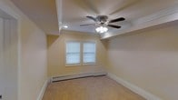 This single-floor space showcases timeless architectural details that contribute to a clean and comfortable living environment. The main room is defined by an elegant tray ceiling with crisp crown molding, adding a sense of depth and refinement to the interior. A multi-blade ceiling fan with an integrated light fixture is centrally mounted, providing both air circulation and ambient lighting. The room is finished with wall-to-wall carpet in a neutral tone, lending a warm and understated feel underfoot. Natural light filters in through two double-hung windows dressed with horizontal blinds, offering both brightness and privacy control. A baseboard heating system runs along the perimeter of the room, ensuring consistent warmth throughout the space. The walls are painted in a soft, neutral hue that complements the overall palette and enhances the sense of openness. Crisp white baseboards frame the floor, adding a polished finishing touch. The space is well-suited for a variety of uses, with a layout that is both practical and inviting. Overall, the interior strikes a balance between classic design elements and functional comfort.