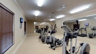 Located at 4500 Highway 57 in Ocean Springs, Mississippi, this fitness center is a well-equipped, single-floor facility designed to support a comprehensive workout experience. The space is organized into distinct cardio and strength-training zones, allowing users to move efficiently between equipment types.

The cardio area features multiple elliptical trainers, treadmills, and stationary bikes arranged in an open layout with generous floor space between machines. A wall-mounted television is positioned for viewing during cardio sessions. The strength-training zone is outfitted with a selection of cable and weight machines suited for a full-body workout routine.

Throughout the facility, large wall mirrors line multiple sides of the room, visually expanding the space and allowing users to monitor their form. The interior is finished with neutral warm-toned walls, crown molding, and light carpet flooring, lending the space a clean and polished aesthetic. Ceiling fans and flush-mounted fluorescent lighting ensure consistent illumination and airflow across the entire area. Windows fitted with dark wood blinds allow for natural light while maintaining privacy.

The overall design is functional yet refined, with a layout that comfortably accommodates multiple users simultaneously. The combination of quality equipment, ample open floor space, and thoughtful finishes makes this fitness center a well-rounded amenity.