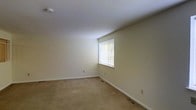 This single-floor residential space presents a straightforward and functional layout with a clean, understated aesthetic throughout. The interior is finished with neutral cream-toned walls that create a bright and open atmosphere, complemented by wall-to-wall carpeting in a warm beige tone that adds a sense of comfort underfoot. The room benefits from a window fitted with vertical blinds, allowing for adjustable natural light to fill the space. Flush-mounted flooring vents indicate a central HVAC system for year-round climate control. The white ceiling is smooth and uniform, contributing to the overall sense of cleanliness and simplicity. White baseboards frame the perimeter of the room, providing a classic finishing touch that ties the neutral palette together. The space is unfurnished, making it easy to envision a variety of furniture arrangements and interior configurations. Electrical outlets are conveniently placed along the walls, offering practical accessibility. Overall, this space is well-suited for those seeking a move-in-ready environment with a clean slate for personalization and customization.