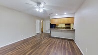 This single-story, one-bedroom, one-bathroom unit offers a functional and efficient layout across a compact footprint. Dark wood-style laminate flooring runs consistently throughout the entire space, creating a cohesive and visually grounding aesthetic. The textured ceiling adds dimension, while neutral light-gray walls provide a clean, versatile backdrop.

The open-concept living area flows directly into the kitchen, maximizing the sense of space. The kitchen features warm honey-toned wood cabinetry, granite-look countertops, black appliances including a full-size refrigerator and built-in microwave, and a raised peninsula that doubles as a breakfast bar. Recessed lighting supplements the natural light that enters through vertical blind-covered sliding doors and windows in the living area.

A ceiling fan with an integrated light fixture serves the living space, contributing to both comfort and illumination. The bedroom is a well-proportioned, quiet room with its own window, ceiling fan, and ample wall space. The full bathroom features a tub-shower combination with white tile surround, a wood-toned vanity with a white countertop, a large mirror, and matching wood-style flooring, maintaining design continuity throughout the unit.