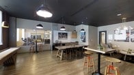 This single-floor amenity space showcases a thoughtfully designed interior that blends industrial aesthetics with modern comfort. The main lounge area is anchored by a large communal dining table with mixed seating, complemented by high-top bar tables and stools that encourage casual gathering and socializing. Striking oversized pendant lights in matte black hang from a dramatic dark ceiling, creating a bold contrast against the light gray walls and warm wood-look flooring that runs throughout the space.

A built-in credenza with wood-paneled cabinetry lines one wall, adding a sleek, functional element to the room. Curated artwork and signage lend a lively, urban character to the space. Natural light filters in through glass partitions, contributing to an open and airy atmosphere.

Adjacent to the lounge, a fully equipped fitness center is visible through floor-to-ceiling glass walls, offering transparency and a seamless connection between the social and wellness areas. The fitness space is outfitted with a range of exercise equipment, making it a practical and accessible amenity.

Overall, the space balances an energetic, contemporary design with versatile functionality — ideal for both active use and relaxed social interaction.