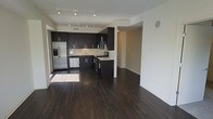 This contemporary single-floor space is defined by its open-concept layout, seamlessly connecting the kitchen and living areas into one cohesive, well-lit environment. Dark hardwood flooring runs throughout, providing a rich, warm contrast to the crisp white walls and ceilings.

The kitchen is a standout feature, outfitted with full-height dark espresso cabinetry offering generous storage, light-toned countertops, and a full suite of stainless steel appliances — including a French door refrigerator with ice and water dispenser, a gas range with overhead microwave, and a dishwasher. A peninsula with additional cabinetry provides extra prep space and subtly defines the kitchen from the open living area beyond.

Track lighting installed along the ceiling efficiently illuminates both the kitchen and the adjoining living space, ensuring consistent brightness throughout. A structural column provides architectural definition between zones without interrupting the open feel of the floor plan.

The living area is expansive and versatile, offering ample room for a variety of furniture arrangements. Clean lines, neutral tones, and quality finishes throughout give this space a polished, modern aesthetic well-suited for both everyday living and entertaining.