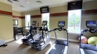 This fitness center offers a dedicated space for cardiovascular exercise, outfitted with professional-grade True-brand equipment including multiple elliptical trainers and a treadmill. The layout is thoughtfully arranged to provide ample room between machines, ensuring a comfortable and safe workout environment. Wall-mounted flat-screen televisions are positioned for easy viewing during exercise sessions, adding a functional entertainment element to the space. Large windows line the walls, flooding the interior with natural light and providing pleasant views of the surrounding greenery and outdoor scenery — creating an inviting atmosphere that encourages activity. The flooring features a combination of hardwood-style and carpeted surfaces, contributing to both durability and comfort underfoot. The walls are painted in a neutral beige tone, accented by a distinctive dark red horizontal stripe that adds a bold design element and visual structure to the room. Recessed lighting in the ceiling complements the natural light provided by the windows. Overall, the fitness center presents a clean, well-maintained environment suited for residents or guests seeking a convenient and fully equipped space for their daily cardio routines.