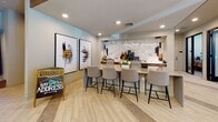 Located at 1000 Artistry Dr in Midlothian, VA, this contemporary single-story community clubhouse showcases a sophisticated blend of modern design and welcoming functionality. The main entry corridor sets the tone immediately, featuring light wood-look flooring with contrasting stone tile inlays, recessed lighting, and a textured stone accent wall that adds warmth and visual depth.

The lounge area is anchored by a tufted navy sofa, mid-century modern accent chairs, and an artfully layered area rug, all set against a whitewashed brick feature wall with subtle LED cove lighting. Large windows invite natural light and views of the surrounding community. An adjacent gathering counter with bar-height seating and bold abstract artwork provides a casual social hub.

A dramatic entertainment room commands attention with bold black-and-white marble-look wall tiles, a sleek linear fireplace, mounted flat-screen displays, a shuffleboard table, and an eye-catching metallic chandelier. Private conference and co-working rooms accessible through floor-to-ceiling glass doors offer a refined setting for focused work or meetings.

A well-appointed bar area features a white quartz countertop, herringbone tile backsplash, pendant lighting, and bar seating, ideal for community gatherings. Soaring ceilings near the rear entrance flood the space with natural light, completing this polished and amenity-rich clubhouse.