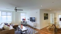 This single-floor space showcases a clean, modern aesthetic defined by crisp white walls, rich hardwood flooring, and a bright, open layout. The living area is the heart of the space, anchored by a sleek white media console and a flat-screen television. Recessed lighting and a contemporary ceiling fan work together to create a well-lit, comfortable atmosphere, complemented by natural light streaming in through a large window. The space is thoughtfully arranged with eclectic furnishings, including a classic tufted armchair, a rustic wood coffee table, and a bold geometric-patterned area rug that adds a lively visual contrast to the neutral backdrop. Decorative accents — including a sculptural deer figurine, colorful throw pillows, and curated wall art — lend personality and warmth to the interior. Wide baseboards and clean architectural lines give the space a polished, contemporary feel throughout. The open flow of the layout promotes a sense of spaciousness, making the area well-suited for both relaxing and entertaining. Overall, the space balances modern design with inviting, lived-in character.