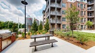 Located at 215 Reformation Pkwy in Canton, Georgia, this outdoor courtyard space serves as a central common area within a multi-story brick residential community. The architecture is defined by classic red brick exteriors accented with arched window details, gray balconies, and dark metal railings that extend across multiple levels, creating a cohesive and attractive streetscape. Wide, smooth concrete walkways wind through the courtyard, providing easy pedestrian access throughout the grounds. Thoughtfully designed landscaping includes neatly arranged shrubs, ornamental grasses, young deciduous trees, and fresh mulched planting beds that add texture and greenery to the space. A standout feature is the expansive open lawn area situated between the building wings, offering open views toward a natural treeline and an open sky, giving the courtyard a spacious, park-like feel. Outdoor seating, including benches and picnic-style tables, is integrated throughout the hardscaped areas, providing comfortable spaces for residents to gather and relax. Decorative street-style light poles are positioned along the walkways to ensure the space remains welcoming after dark. The overall ambiance is clean, modern, and inviting, blending traditional brick craftsmanship with contemporary landscaping design.