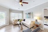 Located at 2151 Cumberland Pkwy SE in Atlanta, GA, this 2-bedroom, 2-bathroom residence offers a thoughtfully designed open-concept layout finished with light hardwood-style flooring and neutral tones throughout. The living room is bright and inviting, featuring crown molding, a ceiling fan, and large windows dressed with sheer curtains that invite abundant natural light and views of the surrounding greenery. A glass door provides direct access to an outdoor balcony area. The dining area is positioned adjacent to the kitchen and living space, illuminated by a modern pendant chandelier. The kitchen is outfitted with white shaker cabinetry, quartz countertops, a grey subway tile backsplash, stainless steel appliances, and a breakfast bar with bar seating. A dedicated in-unit laundry closet with full-size washer and dryer adds everyday convenience. Both bedrooms feature plush carpeting, ceiling fans, walk-in closets with built-in shelving, and generous window coverage for natural light. The primary bedroom includes direct access to its own bathroom, which offers a clean white vanity and a tub-shower combination. The second bathroom mirrors the same refined, functional design. Two dedicated closets provide ample storage throughout this well-organized home.