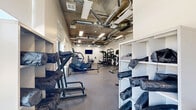 Located at 880 W 200 S in Salt Lake City, Utah, this fitness center offers a comprehensive workout facility designed to support a wide range of exercise routines. The space is housed on a single floor and is thoughtfully organized to maximize usability and flow.

The interior showcases an industrial aesthetic, with exposed concrete ceilings, visible ductwork, and overhead fluorescent lighting that creates a bright, energetic atmosphere. The floors are finished with durable rubber-speckle gym flooring, providing traction and comfort throughout.

The cardio zone features a well-stocked row of treadmills, elliptical machines, and a rowing machine, all positioned along a windowed wall that allows natural light to filter in. The strength training area is generously equipped with multi-tiered dumbbell racks, a cable and pulley machine, a weight bench, and a multi-functional rack system, catering to both beginners and advanced users. Wall-to-wall mirrors in the strength section enhance the sense of space while allowing users to monitor their form.

A dedicated shelving unit stores additional fitness accessories, and a wall-mounted flat-screen TV adds to the environment. A glass-paned entry door offers views to the exterior, further contributing to the open, airy feel of the space. The facility is clean, modern, and well-organized, making it a fully functional fitness destination.