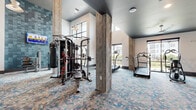 Located in Pooler, GA, this fitness center is a thoughtfully designed, single-level facility that combines modern aesthetics with comprehensive workout capabilities. The space is defined by soaring ceilings that create an open, airy atmosphere, complemented by large windows throughout that invite abundant natural light and provide views of the surrounding exterior.

The interior is unified by a distinctive patterned carpet in earthy tones accented with blue geometric shapes, which adds visual interest while providing a comfortable underfoot surface. Structural columns clad in wood-toned finishes add a warm, contemporary contrast to the crisp white walls and dark ceiling treatments found throughout. A steel-and-wood open staircase serves as a striking architectural feature, reinforcing the modern industrial character of the space.

The fitness area is divided into clearly defined zones. A strength training section houses a multi-functional cable machine, punching bag, adjustable bench, and a mounted television, set against a teal-tiled accent wall. A dedicated free weights area features multiple dumbbell racks alongside full-length mirrors. The cardio zone is well-appointed with treadmills, stair climbers, and cycling equipment. An additional room offers cable machines, rowing, and cycling equipment, with direct access to an outdoor area. Ceiling fans, recessed lighting, and glass-panel doors complete the polished, functional environment.