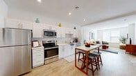This single-floor residence is designed with a clean, contemporary aesthetic, characterized by white walls, recessed lighting, and light-toned hardwood-style flooring that flows seamlessly throughout the main living areas. The open-concept layout connects the kitchen, dining, and living spaces, creating a cohesive and functional environment.

The kitchen is a standout feature, outfitted with crisp white shaker cabinetry extending to the ceiling, a geometric-patterned tile backsplash, light quartz countertops, and a full suite of stainless steel appliances including a refrigerator, range, microwave, and dishwasher. A freestanding dining table with bar stools sits adjacent to the kitchen, serving as a casual dining and gathering area.

The living room offers a comfortable, well-lit space with a large window that brings in natural light. The bedroom features plush carpeting, a large sliding glass door opening to a private balcony with outdoor views, and a direct sightline to the kitchen area. The bathroom is finished with white shaker cabinetry, a undermount sink, quartz countertop, and a framed mirror, maintaining the consistent, modern palette found throughout the residence.