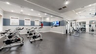 This fitness center offers a comprehensive workout facility thoughtfully organized into distinct exercise zones. The space is finished with durable rubber flooring throughout and features a striking two-tone wall design — a bold slate blue upper band over a light gray lower half — accented by white crown molding and baseboards that lend a polished, cohesive aesthetic.

The main cardio area is generously sized, accommodating a row of spin bikes positioned in front of a wall-mounted flat-screen TV for guided workout sessions. Additional cardio equipment, including treadmills and elliptical machines, occupies a dedicated zone alongside an open stretch of floor ideal for functional training.

A separate strength training area is defined by a textured stone accent column and features a full rack of dumbbells, multiple cable and weight machines, and an adjustable bench. Large windows in this section invite natural light and provide views of the surrounding exterior. A large ceiling fan overhead enhances air circulation during intense workouts.

Full-length mirrors are strategically placed to support proper form and visually expand the space. Practical amenities include a water fountain, waste receptacles, and a fire safety station. Recessed lighting and an exit-lit entryway ensure the space remains bright and safe. The facility is accessible via a glass-paneled entry door, creating a welcoming and functional environment for all fitness levels.