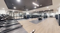 Located at 201 Pilot St in Savannah, Georgia, this contemporary fitness center offers an expansive single-floor layout designed to support a comprehensive workout experience. The space features a sleek, modern aesthetic defined by a striking contrast of dark charcoal ceilings and crisp white walls, accented by light wood-tone flooring and dark rubber mat zones that delineate functional training areas.

The facility is generously outfitted with a wide array of strength training equipment, including cable machines, multi-functional racks, free weight stations with a full dumbbell rack, and various benches. A dedicated cardio zone features a row of TRUE treadmills and rowing machines, ensuring ample options for aerobic training. Large arched mirror panels line the walls, enhancing the sense of openness and allowing users to monitor their form throughout workouts.

Architectural details add a polished, upscale character to the space. Floor-to-ceiling black-framed glass partitions divide areas while maintaining visual connectivity throughout, and recessed lighting provides bright, even illumination across the floor. Ceiling fans and pendant light fixtures contribute both function and style. The entrance area features black-framed glass doors that open to an adjoining stairwell, reflecting a thoughtfully integrated design. Overall, the space presents a refined, high-end fitness environment suited for serious training.