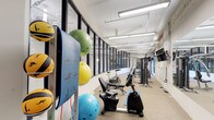 This fitness center is thoughtfully designed to accommodate a wide range of workout styles and fitness goals. The space is defined by its industrial-inspired aesthetic, featuring exposed white-painted brick walls, visible overhead ductwork, and sleek linear fluorescent lighting that ensures the entire area is well illuminated throughout the day.

A striking curved glass partition wall with black steel framing runs along one side of the space, adding a modern architectural element while visually connecting different zones of the facility. Neutral, textured carpet tile flooring provides a comfortable and durable surface underfoot.

The cardio zone is generously stocked with a row of treadmills and elliptical machines, positioned alongside large windows that offer natural light and outside views, creating an energizing environment. A wall-mounted flat-screen television is conveniently placed for entertainment during workouts.

The strength and functional training area features a multi-station cable machine, adjustable weight benches, a rack of dumbbells, and a recumbent exercise bike. A dedicated accessories wall holds an assortment of medicine balls, stability balls in multiple sizes, resistance bands, and exercise mats, making the space versatile for stretching, core work, and group training. The overall ambiance is clean, open, and purposefully organized to support an effective and comfortable fitness experience.