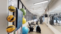 This fitness center is thoughtfully designed to accommodate a wide range of workout styles and fitness goals. The space is defined by its industrial-inspired aesthetic, featuring exposed white-painted brick walls, visible overhead ductwork, and sleek linear fluorescent lighting that ensures the entire area is well illuminated throughout the day.

A striking curved glass partition wall with black steel framing runs along one side of the space, adding a modern architectural element while visually connecting different zones of the facility. Neutral, textured carpet tile flooring provides a comfortable and durable surface underfoot.

The cardio zone is generously stocked with a row of treadmills and elliptical machines, positioned alongside large windows that offer natural light and outside views, creating an energizing environment. A wall-mounted flat-screen television is conveniently placed for entertainment during workouts.

The strength and functional training area features a multi-station cable machine, adjustable weight benches, a rack of dumbbells, and a recumbent exercise bike. A dedicated accessories wall holds an assortment of medicine balls, stability balls in multiple sizes, resistance bands, and exercise mats, making the space versatile for stretching, core work, and group training. The overall ambiance is clean, open, and purposefully organized to support an effective and comfortable fitness experience.