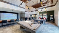 Located at 4900 East Deer Valley Drive in Phoenix, AZ, this expansive game room is a premier entertainment destination designed for socializing and recreation. The space is defined by its dramatic soaring ceilings adorned with elegant wood slat ceiling details and striking sculptural pendant lighting, creating a bold architectural statement throughout.

The room is thoughtfully organized into distinct activity zones. Multiple pool tables serve as centerpieces, surrounded by open floor space finished with patterned carpet tiles. An arcade section features classic pinball and racing game machines, while a dedicated golf simulator bay offers an immersive experience, partially enclosed by a textured metal mesh partition for ambiance and separation.

A generous lounge and bar seating area includes a tufted banquette, high-top tables, and chrome bar stools, complemented by stylish wall sconces. A projection screen and mounted flat-screen television provide additional entertainment options. A shuffleboard table adds further variety to the gaming offerings.

Large floor-to-ceiling windows flood the space with natural light and offer views of the surrounding Phoenix streetscape and palm trees. Black-framed glass doors connect seamlessly to adjacent common areas, enhancing the open, sociable atmosphere. The overall design blends modern industrial aesthetics with warm wood accents, delivering a dynamic and inviting environment.