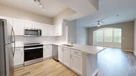 Located at 1000 Fanfare Way in Alpharetta, Georgia, this single-story residence offers a thoughtfully designed open-concept layout with modern finishes throughout. The kitchen is a standout feature, showcasing white shaker cabinetry, a classic white subway tile backsplash, quartz countertops, and a peninsula with a built-in sink that overlooks the adjoining living area. Stainless steel appliances, including a range, microwave, dishwasher, and refrigerator, complete the well-appointed space. Light wood-look flooring flows seamlessly through the kitchen and main living areas, lending a warm and cohesive aesthetic. The spacious living room features built-in shelving, a ceiling fan, and an arched entryway leading to a bright nook with large windows that frame lush greenery outside, filling the space with natural light. The bedroom is generously sized, finished with plush carpet, a ceiling fan, and a large window offering additional garden views. The primary bathroom includes a walk-in closet with wire shelving and an accessible toilet area with a grab bar. Neutral wall tones and clean white trim throughout create a bright, move-in-ready atmosphere that is both functional and inviting.