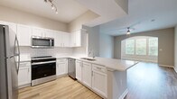 Located at 1000 Fanfare Way in Alpharetta, Georgia, this single-story residence offers a thoughtfully designed open-concept layout with modern finishes throughout. The kitchen is a standout feature, showcasing white shaker cabinetry, a classic white subway tile backsplash, quartz countertops, and a peninsula with a built-in sink that overlooks the adjoining living area. Stainless steel appliances, including a range, microwave, dishwasher, and refrigerator, complete the well-appointed space. Light wood-look flooring flows seamlessly through the kitchen and main living areas, lending a warm and cohesive aesthetic. The spacious living room features built-in shelving, a ceiling fan, and an arched entryway leading to a bright nook with large windows that frame lush greenery outside, filling the space with natural light. The bedroom is generously sized, finished with plush carpet, a ceiling fan, and a large window offering additional garden views. The primary bathroom includes a walk-in closet with wire shelving and an accessible toilet area with a grab bar. Neutral wall tones and clean white trim throughout create a bright, move-in-ready atmosphere that is both functional and inviting.
