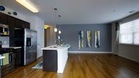 This single-floor space showcases a contemporary open-concept layout that seamlessly connects the kitchen and living areas. The kitchen is anchored by a sleek, curved island with a white countertop, an undermount sink, and a modern arc faucet. Dark cabinetry on the island provides a striking contrast against the bright white surface, while stainless steel appliances, including a full-size refrigerator, add a professional touch. Two pendant lights with frosted globe shades hang above the island, providing focused task lighting and contributing to the modern aesthetic. Warm-toned hardwood flooring extends throughout the space, adding richness and continuity to the open floor plan. The walls are finished in a cool, sophisticated grey tone that complements the overall color scheme. The living area features a generous open wall adorned with a trio of abstract canvas artworks in blue, white, and gold, serving as a focal point. A doorway visible in the background hints at additional rooms, and the overall layout offers a bright, well-organized living environment with a clean, modern design sensibility.