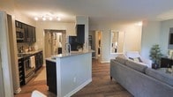 This single-floor residence offers a thoughtfully designed open-concept layout that connects the kitchen, dining, and living areas in a cohesive, functional arrangement. The kitchen serves as a central focal point, featuring a peninsula with a granite countertop that provides both prep space and a casual seating area. Dark cabinetry contrasts elegantly with the light granite surface, while a stainless steel faucet adds a modern finishing touch. Track lighting overhead ensures the kitchen remains well-lit and practical.

Rich hardwood-style flooring flows throughout the main living area, lending warmth and visual continuity to the space. The open living area is generously sized, accommodating comfortable seating while maintaining an airy, uncluttered feel. Neutral gray walls create a versatile backdrop that complements the darker design elements throughout.

Hallways branch off from the main living area, leading to additional rooms including a bathroom and bedroom, both of which are visible in the background. A large full-length mirror in the living area enhances the sense of space and light. The overall ambiance is modern and cohesive, with a clean aesthetic that balances style and everyday functionality throughout the home.