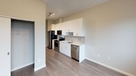 This single-floor unit offers a clean, contemporary design with cohesive finishes throughout. The open-concept layout connects the kitchen and living area, creating a functional and visually spacious environment. Wood-look flooring runs throughout the unit, providing a warm, consistent aesthetic.

The kitchen is a standout feature, outfitted with white flat-front cabinetry extending to the ceiling for ample storage, white quartz countertops, and a herringbone-patterned tile backsplash. A full suite of stainless steel appliances includes a refrigerator, electric range, over-the-range microwave, and dishwasher. Track lighting overhead illuminates the workspace efficiently.

The living area is open and well-proportioned, benefiting from natural light and direct sightlines into the kitchen. A closet is accessible from this shared space, adding practical storage.

The bedroom features two windows with views of the surrounding neighborhood's tree canopy, allowing natural light and outdoor scenery to fill the room. A wall-mounted HVAC unit provides climate control.

The bathroom is finished with a white vanity featuring ample cabinetry, a framed mirror with overhead lighting, white quartz countertop, and a full tub and shower combination — all consistent with the unit's modern, neutral palette.