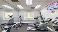 Located at Arbor Crossing in Lithonia, Georgia, this fitness center offers a practical and well-organized exercise space designed to support a range of cardio workouts. The room is finished with neutral gray walls accented by white chair-rail molding, dark low-pile carpet flooring, and a drop tile ceiling with recessed fluorescent lighting that keeps the space bright and functional throughout the day.

A large wall-mounted mirror spans a significant portion of the room, visually expanding the space and allowing users to monitor their form during workouts. The equipment lineup includes multiple TRUE-brand treadmills, elliptical trainers, and stationary bikes — including a recumbent model — providing a variety of cardio options. A wall-mounted flat-screen television adds an entertainment element for those longer sessions.

A window overlooking what appears to be a pool area introduces natural light and an outdoor view, enhancing the overall atmosphere. Two white modern accent chairs are positioned near the window, offering a casual seating area. A hand sanitizer dispenser is mounted on the wall, and a fire extinguisher is visibly stationed for safety compliance. Two access doors provide convenient entry and exit. Overall, the space is clean, efficiently arranged, and well-suited for daily cardiovascular exercise.