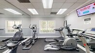 Located at Arbor Crossing in Lithonia, Georgia, this fitness center offers a practical and well-organized exercise space designed to support a range of cardio workouts. The room is finished with neutral gray walls accented by white chair-rail molding, dark low-pile carpet flooring, and a drop tile ceiling with recessed fluorescent lighting that keeps the space bright and functional throughout the day.

A large wall-mounted mirror spans a significant portion of the room, visually expanding the space and allowing users to monitor their form during workouts. The equipment lineup includes multiple TRUE-brand treadmills, elliptical trainers, and stationary bikes — including a recumbent model — providing a variety of cardio options. A wall-mounted flat-screen television adds an entertainment element for those longer sessions.

A window overlooking what appears to be a pool area introduces natural light and an outdoor view, enhancing the overall atmosphere. Two white modern accent chairs are positioned near the window, offering a casual seating area. A hand sanitizer dispenser is mounted on the wall, and a fire extinguisher is visibly stationed for safety compliance. Two access doors provide convenient entry and exit. Overall, the space is clean, efficiently arranged, and well-suited for daily cardiovascular exercise.