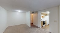 Located at 1051-1057 Broadway in Chula Vista, this single-story unit offers a straightforward and functional layout designed for comfortable living. The main living area is finished with wall-to-wall carpeting in a warm neutral tone, complemented by crisp white walls and a textured ceiling. Overhead lighting provides consistent illumination throughout the space, while HVAC vents indicate a climate control system integrated into the ceiling.

The kitchen is open and directly accessible from the living area, creating a connected feel between the two spaces. It features light wood cabinetry offering ample storage, a white electric range with a stainless steel finish, and an overhead ventilation hood. The kitchen is finished with hard-surface flooring, contrasting with the carpeted living space.

A hallway visible from the main living area leads to additional rooms, suggesting a clear separation between living and private spaces. The overall interior maintains a clean, neutral aesthetic with a practical design focus, making it a versatile and move-in-ready space in the Chula Vista community.