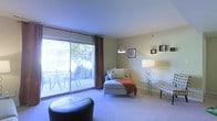 This single-floor residence features a welcoming living area defined by soft, neutral-toned walls and plush carpeted flooring that adds warmth underfoot. The room is anchored by a large sliding glass door dressed with rich brown curtains, which floods the space with abundant natural light and provides direct access to a private outdoor patio, where patio furniture is visible surrounded by lush greenery.

The living area is thoughtfully arranged with a cushioned chaise lounge accented by decorative pillows and a warm orange throw blanket, creating a comfortable and inviting focal point. A sleek glass side table paired with an elegant double-globe table lamp adds a touch of modern sophistication. Decorative wall-mounted shelving units and framed artwork contribute personality and visual interest to the walls.

An arc floor lamp with a bowl-shaped shade provides additional ambient lighting, while a tufted leather ottoman positioned in the foreground offers versatile functionality. The overall ambiance of the space is calm and airy, blending functional furnishings with tasteful décor to create a cohesive, livable environment with a seamless connection to the outdoors.