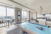 This 1,400 square foot clubroom is a thoughtfully designed amenity space spanning a single floor, offering residents a sophisticated environment for relaxation and socializing. The lounge area is anchored by a modern fireplace with a wall-mounted TV above, complemented by rich leather armchairs, a coffee table, and warm hardwood flooring. Floor-to-ceiling glass windows and sliding doors flood the space with abundant natural light while providing stunning panoramic city skyline views, creating an elevated, airy atmosphere throughout.

A fully equipped kitchen features a large white quartz island with bar seating, stainless steel appliances including a double wall oven and gas cooktop, dark cabinetry, and stylish pendant lighting. Adjacent to the kitchen, a dedicated workspace with built-in desk and cabinetry offers a quiet area for focused tasks.

The game area is highlighted by a full-size billiards table set against large glass walls and sliding doors that open directly to an outdoor terrace, seamlessly blending indoor and outdoor living. High ceilings with recessed lighting, tray ceiling details, and a neutral palette of whites and grays with teal accent walls contribute to a modern, upscale ambiance throughout the space.