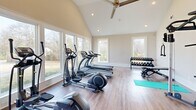 Located in Grovetown, GA, this fitness center offers a bright, welcoming environment designed to support a well-rounded workout routine. The space is defined by its striking vaulted ceilings, which lend an open and airy feel throughout. Large windows line the walls, flooding the room with natural light and providing pleasant views of the surrounding community and greenery, creating an energizing atmosphere for exercise.

The flooring throughout is a warm-toned hardwood-style surface, adding a clean and polished aesthetic while remaining durable underfoot. A ceiling fan keeps air circulating, complementing the recessed lighting overhead for comfortable use at any time of day.

Cardio equipment takes center stage, with multiple treadmills and elliptical machines positioned to take advantage of the expansive window views. A stationary bike and rowing machine round out the cardiovascular offerings. Strength training is well-represented with a multi-functional cable machine, a full rack of dumbbells, and a weight bench for targeted resistance work. Exercise accessories including medicine balls, stability balls, and yoga mats provide additional versatility for stretching, balance, and functional fitness routines.

The layout is thoughtfully organized to allow generous space between equipment, enabling comfortable movement throughout. A posted fitness center rules sign reflects a community-oriented facility, making this a practical and inviting space for residents to pursue their fitness goals.