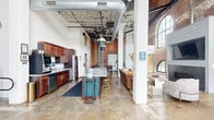 Located at 1101 Hawthorne Lane in Charlotte's vibrant Plaza Midwood district, this expansive two-story space masterfully blends historic industrial character with modern amenities. The building's original architecture is celebrated throughout, with exposed brick walls, structural steel beams, and polished concrete floors creating a striking industrial-chic aesthetic across both levels.

The ground floor commands attention with its soaring ceiling height, anchored by a dramatic living area featuring a herringbone-tiled fireplace framed by arched brick windows that flood the space with natural light. Adjacent to this is a fully equipped kitchen with rich wood cabinetry, granite countertops, a statement island with geometric patterned panels, and stainless steel appliances. The open layout flows seamlessly into a recreation area outfitted with a ping pong table and dartboard, making it ideal for social gatherings.

A bold steel staircase with mesh railings connects to the upper level, where a mezzanine workspace overlooks the main floor below. Large arched windows line the upper level, offering generous natural light and outdoor views. Throughout, Edison-style pendant lighting and string lights complement the raw industrial elements, while carefully curated furnishings and greenery add warmth to the commanding space.