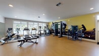 This fitness center at 13420 NE Leroy Haagen Memorial Dr in Vancouver, WA offers a comprehensive, single-level workout facility designed to support a wide range of exercise routines. The space features light hardwood-style flooring throughout, recessed lighting, and a clean white ceiling that keeps the environment bright and open.

Along one wall, a bank of large windows allows generous natural light to fill the room, providing views of the outdoor pool area and greenery beyond. The cardio zone is well-stocked with multiple treadmills, elliptical trainers, and a stair climber, all positioned to take advantage of the natural light. Adjacent to this area, a cable machine and leg press provide dedicated strength training options.

A full-length mirror spans one wall, visually expanding the space and aiding in form correction during workouts. A neatly organized dumbbell rack offers a range of free weights, complemented by flat weight benches positioned throughout the open floor area. A wall-mounted flat-screen TV, exercise balls, and resistance equipment add versatility for functional training.

The standout design feature is a bold, large-scale athletic mural rendered in black silhouette against a yellow backdrop, energizing the atmosphere. French doors connect the fitness center to an adjacent lounge or common area, enhancing the sense of openness and connectivity within the facility.