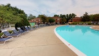 Carlton Apartments offers a welcoming community environment highlighted by a standout outdoor aquatic amenity. The centerpiece of the outdoor space is a large, kidney-shaped swimming pool filled with clear, blue water, designed to accommodate residents looking to relax and recreate. Surrounding the pool is a wide, stamped concrete deck that provides ample room for lounging and socializing. A generous row of pool chairs lines the deck, offering plenty of seating for sunbathing and leisure. The pool area is framed by mature trees that provide natural shade and greenery, enhancing the peaceful, resort-like atmosphere. In the background, the classic brick-exterior residential buildings rise with traditional architectural charm, featuring pitched rooflines and a well-maintained appearance that reflects the community's established character. The lush landscaping throughout the grounds contributes to a serene and inviting outdoor environment. Overall, the Carlton Apartments community combines practical amenities with comfortable, classic architecture to create a welcoming residential setting suited for everyday living.