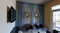 This refined single-floor space makes a striking first impression through its carefully curated interior design. The lounge area serves as a centerpiece, showcasing dramatic floor-to-ceiling steel-blue drapes that lend an air of grandeur and sophistication. Adorning the draped wall are two eye-catching starburst mirrors with ornate metallic frames, serving as bold decorative focal points that reflect light and add a glamorous, artistic touch to the space. The seating arrangement features sleek, dark upholstered armchairs paired with a polished circular metal coffee table, creating an intimate yet stylish conversation area. The flooring is finished in light-toned tile, contributing to a clean and elegant aesthetic throughout. A flat-screen television mounted to one side offers a functional element without disrupting the overall design harmony. The warm yellow-toned walls contrast beautifully with the cool blue drapery, resulting in a balanced and visually engaging color palette. The overall ambiance is one of understated luxury — modern in form, yet rich in decorative detail — making this space well-suited for both relaxed gatherings and more formal occasions.