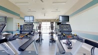 This fitness center is designed to support a well-rounded cardio workout experience in a clean, functional environment. The space is arranged with rows of Matrix-brand cardio equipment, including treadmills and elliptical trainers, each outfitted with individual flat-screen monitors for entertainment during workouts. The layout is symmetrical and spacious, allowing comfortable movement between machines. Light-colored flooring and pale walls trimmed with a soft teal accent band contribute to a bright, airy atmosphere. Ceiling fans are installed overhead, promoting air circulation throughout the room. A large garage-style window at the far end of the space floods the interior with natural light and provides an open, welcoming feel. Mirrored wall panels along the sides enhance the sense of depth and space while allowing users to monitor their form. The overall ambiance is clean and modern, reflecting a well-maintained facility suited for daily fitness use.