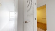 This contemporary single-floor residence showcases a thoughtfully designed layout with clean architectural lines and high-quality finishes throughout. The modern kitchen is a standout feature, equipped with high-gloss white upper cabinetry, rich dark wood lower cabinets, a white glass backsplash, and a full suite of stainless steel appliances including a Bosch wall oven, gas cooktop, and a bottom-freezer refrigerator. Light wood-tone flooring runs seamlessly throughout the living and bedroom areas, lending the space a bright, airy feel. Louvered interior doors add a subtle architectural detail, providing both ventilation and a clean aesthetic. The bathroom is elegantly appointed with large-format marble-look tiles lining both the walls and the soaking tub surround, complemented by warm beige floor tiles and a bold yellow accent wall that introduces a pop of color. A second bathroom area is also present, finished with coordinating tile work. Large floor-to-ceiling windows flood the interior with natural light and frame impressive panoramic views of the surrounding urban skyline, with city buildings and construction cranes visible in the distance, suggesting an elevated position within a dynamic city setting. The overall ambiance is modern, clean, and sophisticated.