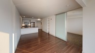 Located at 606 Burwell St, Unit 408 in Bremerton, Washington, this modern single-floor unit offers a clean, contemporary design with an open and airy feel. Rich hardwood flooring flows throughout the main living area, complementing the bright white walls and crisp clean lines that define the space.

The expansive living area is bathed in natural light, courtesy of large sliding glass doors that open onto a private balcony, offering views of the surrounding Bremerton cityscape. A sleek frosted-glass sliding panel provides flexible room division, adding a stylish architectural element while maintaining an open, versatile layout. Built-in shelving adds practical storage without sacrificing the unit's minimalist aesthetic.

The well-appointed kitchen features stainless steel appliances, including a refrigerator, range, and microwave, along with white cabinetry, a dark countertop island, and an undermount sink — ideal for both everyday cooking and entertaining. The entry area provides a smooth transition into the living and kitchen spaces.

The bathroom is finished in a clean, modern style with wood-tone cabinetry, a white countertop vanity, a large mirror, and hardwood-style flooring that ties seamlessly into the rest of the unit. The overall ambiance is bright, fresh, and thoughtfully designed for contemporary urban living.