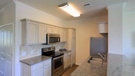 This single-floor residence offers a thoughtfully designed layout that balances functionality and comfort. The kitchen serves as a standout feature, showcasing granite countertops with warm neutral tones that complement the classic white cabinetry lining the walls. A full suite of stainless steel appliances — including an electric range, over-the-range microwave, and refrigerator — provides a modern, cohesive look. A stainless steel sink is conveniently positioned along the countertop, adding to the kitchen's practical design. The cabinetry extends to ceiling height in sections, offering generous storage capacity. A diagonal tile backsplash adds a subtle decorative element that ties the space together. Crown molding trims the ceiling throughout, lending a refined architectural detail to the interior. The flooring transitions to hardwood-style material, contributing warmth and continuity to the space. Overhead flush-mount lighting ensures the kitchen is well-illuminated. The overall ambiance is clean, bright, and move-in ready, with a neutral color palette that creates a welcoming and versatile environment suitable for a variety of interior styles.