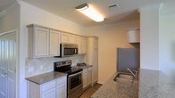 This single-floor residence offers a thoughtfully designed layout that balances functionality and comfort. The kitchen serves as a standout feature, showcasing granite countertops with warm neutral tones that complement the classic white cabinetry lining the walls. A full suite of stainless steel appliances — including an electric range, over-the-range microwave, and refrigerator — provides a modern, cohesive look. A stainless steel sink is conveniently positioned along the countertop, adding to the kitchen's practical design. The cabinetry extends to ceiling height in sections, offering generous storage capacity. A diagonal tile backsplash adds a subtle decorative element that ties the space together. Crown molding trims the ceiling throughout, lending a refined architectural detail to the interior. The flooring transitions to hardwood-style material, contributing warmth and continuity to the space. Overhead flush-mount lighting ensures the kitchen is well-illuminated. The overall ambiance is clean, bright, and move-in ready, with a neutral color palette that creates a welcoming and versatile environment suitable for a variety of interior styles.