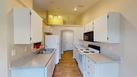 Located at 2415 N Cherry St in Spokane Valley, WA, this single-floor unit offers a well-organized, open-concept layout designed for comfortable everyday living. The galley-style kitchen is efficiently arranged with ample white cabinetry, light granite-style countertops, wood-look flooring, a double-basin sink, and a full suite of appliances including a refrigerator, electric range, dishwasher, and over-the-range microwave. Vaulted ceiling accents above the kitchen add visual interest and an airy feel to the space. A pass-through opening connects the kitchen to the dining area, which features a pendant light fixture and a window that brings in natural light. The living room is generously proportioned with plush carpeting, vertical blinds covering a sliding glass door, and a wall-mounted air conditioning unit. Bedrooms are carpeted and feature neutral wall tones alongside sliding wood-panel closet doors offering practical storage. The bathroom includes wood-look flooring, a full soaking tub with shower, and clean, neutral finishes throughout. The unit's entry is accessed through an arched doorway, adding a subtle architectural touch to the overall design.