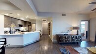 This contemporary single-floor residence showcases a thoughtfully designed open-concept layout that seamlessly connects the kitchen, dining, and living areas. Dark hardwood flooring runs throughout, providing a warm and cohesive foundation that anchors each space.

The kitchen is outfitted with stainless steel appliances, dark wood cabinetry, and a bright white countertop peninsula that doubles as a casual dining surface. Recessed lighting and track lighting illuminate the workspace efficiently, while the open design allows for easy interaction with adjacent living areas.

The living area makes a bold statement with a deep plum accent wall adorned with a striking large-scale artwork, creating a dramatic focal point. A tufted velvet sofa in warm gold anchors the seating arrangement, complemented by a geometric coffee table that adds an artistic touch to the space.

The dining area bridges the kitchen and living room with a defined yet open feel, featuring a stylish dining set beneath soft overhead lighting and a subtle blue-toned accent wall.

A clean, well-lit hallway leads to the entry door, maintaining a bright and welcoming atmosphere. Recessed ceiling lights and modern HVAC vents are integrated discreetly throughout, ensuring comfort without compromising the sleek aesthetic. The overall ambiance is sophisticated, urban, and curated.