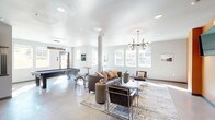 This single-floor community lounge is designed for social interaction and recreation, offering a thoughtfully arranged open-plan layout with distinct activity zones that flow seamlessly into one another. The main lounge area is anchored by a rich brown leather sofa accented with colorful throw pillows, complemented by sleek armchairs and a central coffee table arranged atop a textured area rug. A striking multi-bulb Edison chandelier serves as a focal point overhead, casting warm ambient light across the polished concrete floors. Accent walls in a bold terracotta orange add warmth and visual depth to the otherwise neutral gray and white palette. A wall-mounted flat-screen television provides an entertainment focal point in the seating area. Adjacent to the lounge, a dedicated game room features a full-size pool table positioned beneath a pendant light, with large windows allowing generous natural light and views to an outdoor fence-lined area. A high-top table with bar stools provides additional casual seating. The kitchenette is equipped with dark wood cabinetry, white countertops, a stainless steel sink, and a full-size refrigerator, making the space well-suited for gatherings and events. Throughout the space, recessed lighting and large windows ensure a bright, welcoming atmosphere ideal for community engagement.